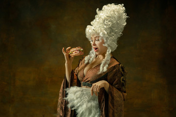 Eating burger. Portrait of medieval young woman in brown vintage clothing on dark background....
