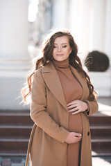 Smiling happy pregnant woman 25-26 year old holding with hands her belly wearing stylish beige jacket outdoors in city street. Motherhood. 20s. Spring season.