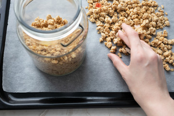 Woman hand poured granola in glass jar on parchment paper on baking sheet, close-up. Made homemade healthy breakfast