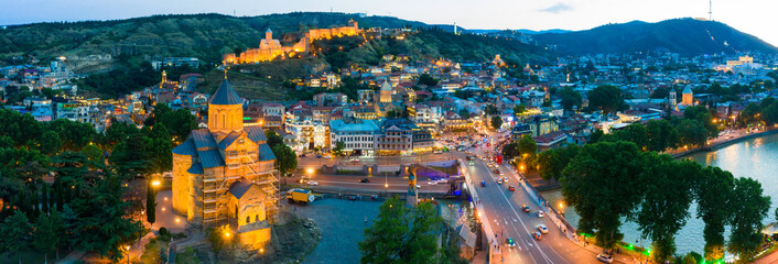 The evening panorama of the old town in the old district of Avlabari, Holy Trinity Cathedral and Rike Park, the Kura river reflects the evening city lights in Tbilisi, Georgia. - obrazy, fototapety, plakaty
