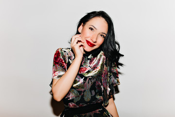 Glamorous black-haired woman with bright makeup posing with surprised smile. Glad female model with dark hair touching her face and laughing on white background.