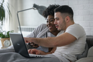 Young smiling multi ethnic couple shopping over internet in the bed in the morning. E-banking and online shopping concept