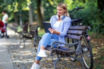 Blonde woman sitting on bench with tablet on her hands and headphones over her neck