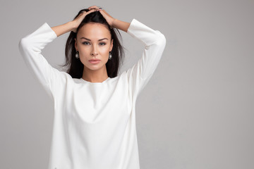 Portrait of casual young brunette woman touching hair by hand looking at camera medium shot. Fashion girl having calmness emotion standing isolated at white background