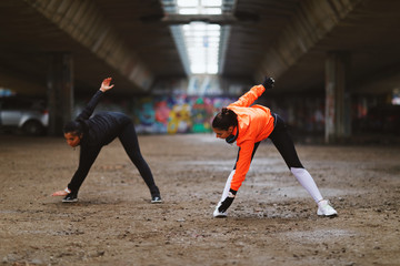 Two young fitness woman runner stretching legs before run under overpasses during a cold day