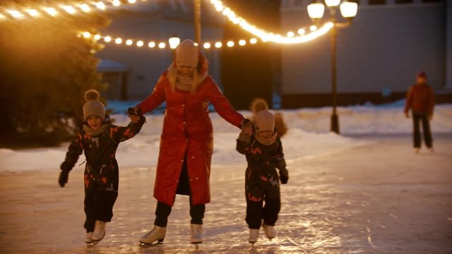 A family of mother and two kids skating on public ice rink at evening in bright lights