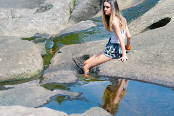 Attractive teenage girl sitting on rock with legs over edge in water.