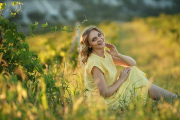 Beautiful young woman in yellow dress sitting on green grass in field.