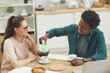 African young man sitting at the table and pouring coffee into the cup of his girlfriend while they sitting in the kitchen