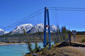 Blue suspension bridge over turquoise river. High mountains covered with snow, blue sky, sunny day. Kootenay plains Ecological Reserve,  Canada.