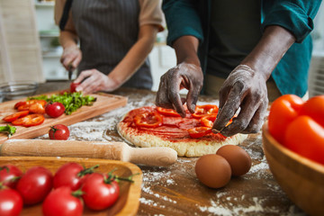 Close-up of African man putting pepper on dough for future pizza with woman cutting vegetables in the background