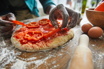 Close-up of African man putting pepper on dough and making vegetable pizza for dinner at the table