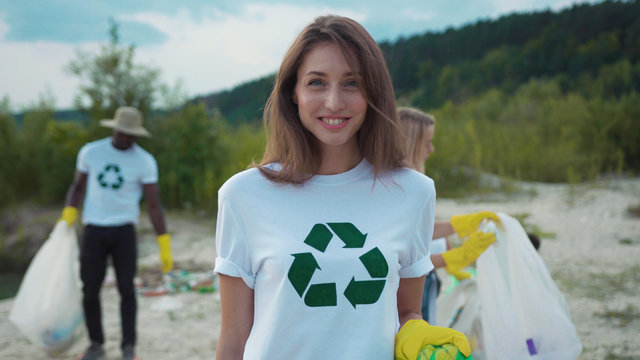 Pretty caucasian girl picking up garbage at sandy beach. Portrait of smiling female eco fighter helping environment cleaning nature from pollution.