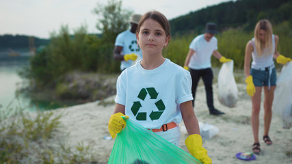 Porrtrait of little cute cheerful girl cleaning the sandy beach from garbage smiling of joy having good time outdoor. Young volunteer team on eco-friendly movement.
