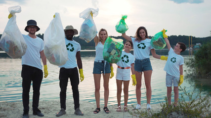 Obraz premium Team of multi-race happy ecology volunteers smiling posing on sandy beach at beautiful lake demonstrating trash bags. Successful nature cleanup. Fight pollution.