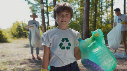 Happy boy voluteers activists child in gloves tidying up rubbish in park or forest look at camera...