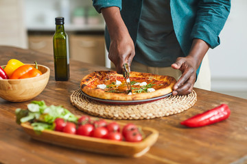 Close-up of African young man standing near the wooden table and cutting pizza with cheese before eating it