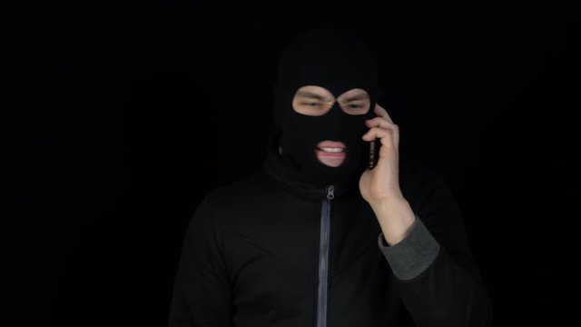 A man in a balaclava mask is talking on a phone. A thug screaming on the phone on a black background.