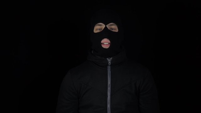 A man in a balaclava mask is standing with a bundle of rubles. The thug leafing through the money and rejoices. On a black background.