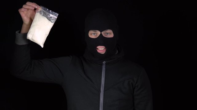 A man in a balaclava mask stands with a pack of heroin. The bandit holds a drug bag and rejoices. On a black background.
