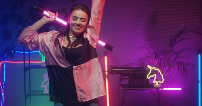 Pretty Caucasian brunette girl dancing cheerfully with astera LED lamp in hands. Beautiful young female having fun in futuristic room with blue and pinh neon light.