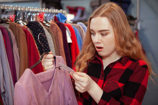 Young woman looking shocked, checking the price of a dress at fashion boutique. Overwhelmed woman looking at price tag of an expensive dress