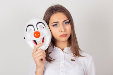 Wearing a mask, faking it concept. worried girl with sad expression holding a clown mask expressing happiness, looking camera, isolated white background. Bipolar. Facial expression feeling