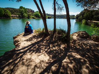 Mujer con camiseta azul y sombrero sentada al borde de la laguna entre la sombra de los arboles