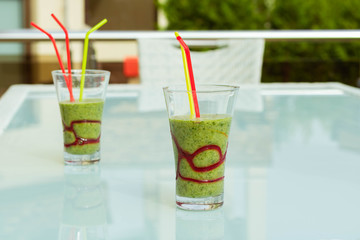 Two three Healthy green smoothie made from spinach, kiwi, bananas and cucumber with green straws on white table on the outdoor veranda