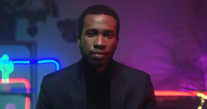 Portrait of handsome African American young man in black looking in camera in room with neon light. Close up of serious good looking stylish guy in club.
