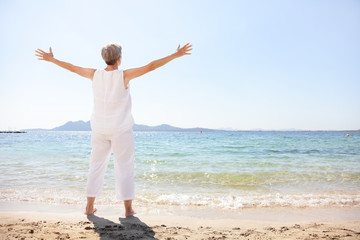 Happy senior woman enjoying freedom of early retirement carefree on summer holiday in happiness standing with arms outstretched at beach. Full length view of lady tourist wearing linen clothing.