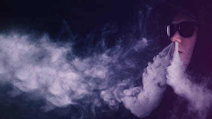 vape man. Portrait of a young white man wearing glasses and a cap lets off steam from an electronic...