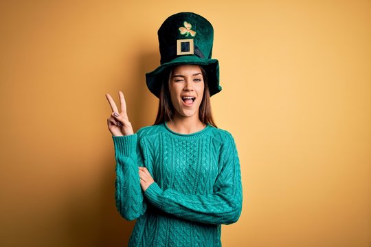 Young beautiful brunette woman wearing green hat with clover celebrating saint patricks day smiling with happy face winking at the camera doing victory sign. Number two.