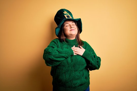 Young beautiful plus size woman wearing green hat with clover celebrating saint patricks day smiling with hands on chest with closed eyes and grateful gesture on face. Health concept.
