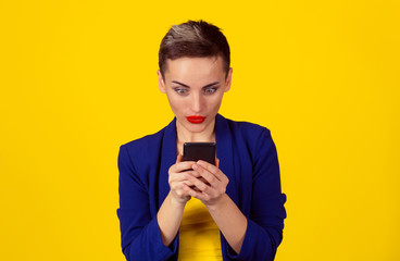 Stunned by sms. Portrait surprised woman typing on smart phone by news saw on cell mobile phone. Multicultural Latina girl short hair red lips blue suit yellow shirt isolated yellow background wall