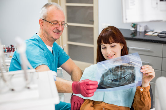 Handsome senior man dentist looking at x-ray image of his young smiling woman patient.