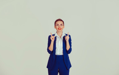 Look up here. Business woman presenting showing copy space above head with index fingers hands isolated light green white office wall background. Short hair red lips blue suit white shirt formal wear