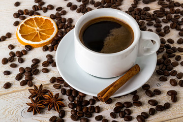 Hot coffee in white elegant cup with saucer on a background of coffe beans over a white wooden table. Coffee with cinnamon, anise and dry citrus.