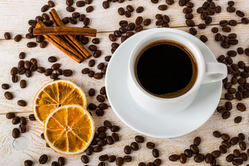 Top view of hot coffee in white elegant cup with saucer on a background of coffe beans over a white wooden table. Coffee with cinnamon, anise and dry citrus.