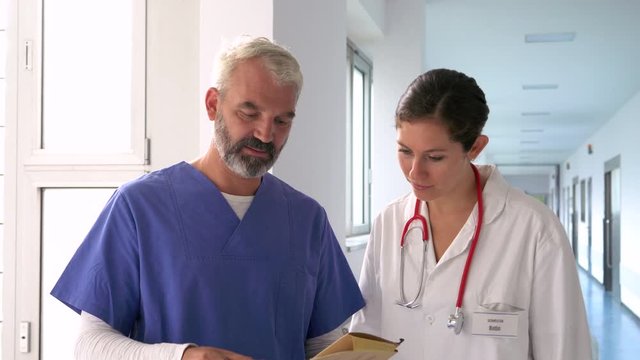 Handheld shot of two doctors greeting and discussing medical record in hospital corridor