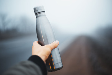 Close-up of male hand holding reusable steel thermo water bottle on background of foggy road.