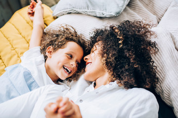 Cheerful young ethnic mother and little daughter having fun on bed at home