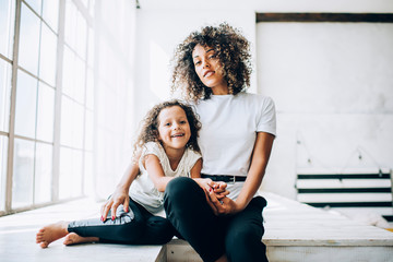 Black mother and daughter sitting near window happily laughing and looking at camera