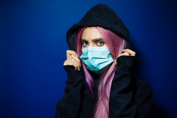 Studio portrait of young girl with pink hair and blue eyes, wearing medical flu mask in hoodie sweater on background of dark blue color.