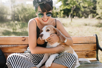 Close-up outdoor portrait of fascinating laughing girl holding beagle puppy while sitting on bench....