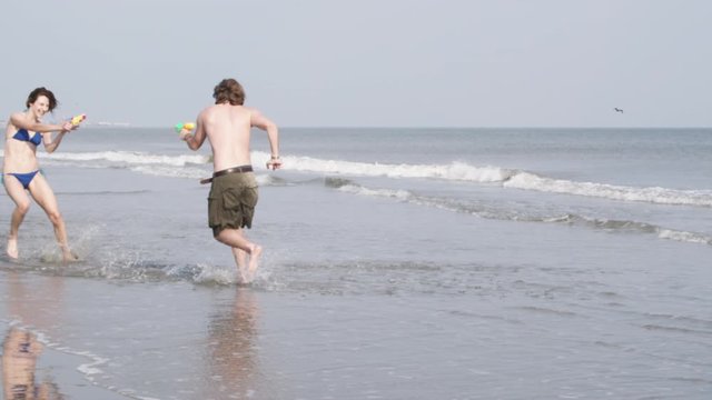 SLO MO, WS, PAN of a young couple playing with squirt guns at the beach