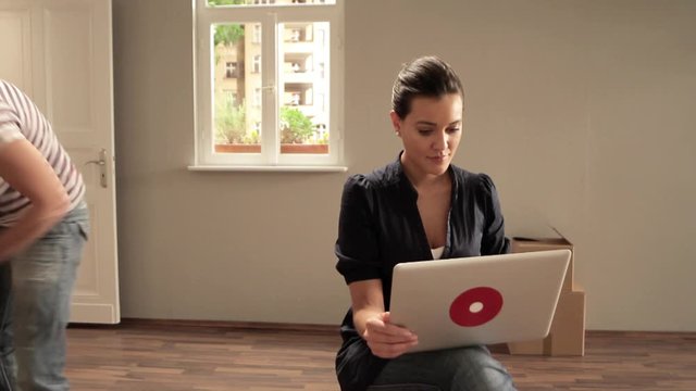 T/L, MS, PAN of a woman using a laptop while movers carry boxes in background