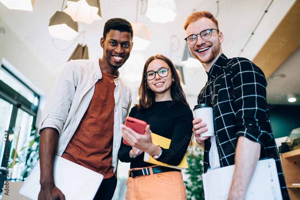 Poster half length portrait of cheerful multicultural classmates standing indoors and smiling at camera dur