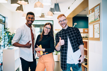 Obraz premium Half length portrait of cheerful diverse male and female hipster standing in public place and smiling at camera feeling prosperous indoors, toothy multicultural students showing OK while posing