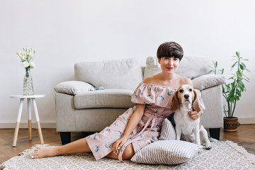 Obraz premium Blissful brunette girl in cute outfit sits on carpet in front of gray sofa with her puppy. Indoor portrait of smiling dark-haired lady and white beagle dog with brown ears posing in room with plant.